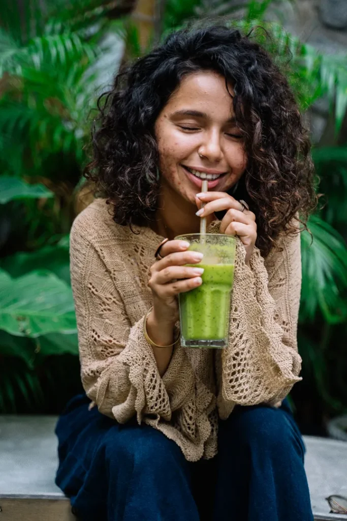 A lady smiling while drinking a green smoothie through a straw while wearing invisible aligners, sitting in front of lush green plants.