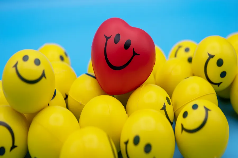 A group of yellow balls with smiley faces are piled together and a red heart shaped ball on a blue background