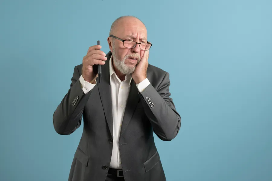 An elderly man with glasses, dressed in a suit, is holding his cheek with one hand and a phone near his ear with the other