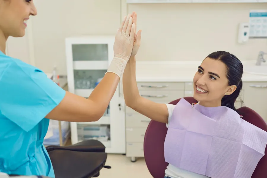 A dental patient sitting in a chair smiles and gives a high-five to a dental professional wearing scrubs and gloves in a clinic