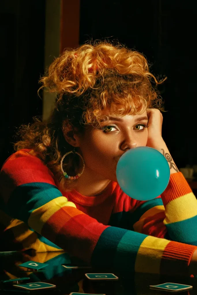 A person with curly hair wearing a striped shirt and hoop earrings blows a bubble with gum while resting their head on one hand