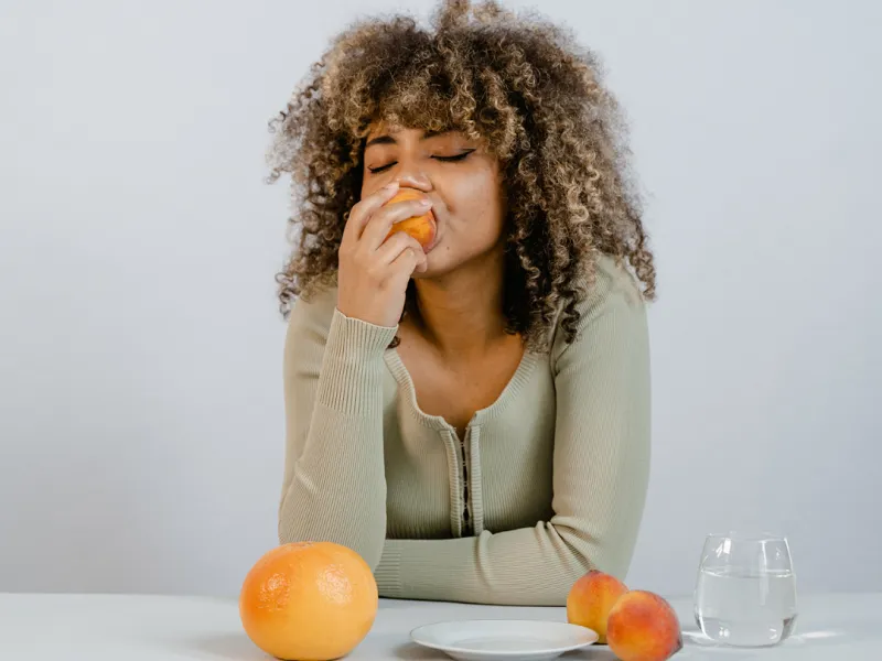 A woman with curly hair and a green sweater eats a peach at a table with another peach, a grapefruit, an empty plate, and a glass of water