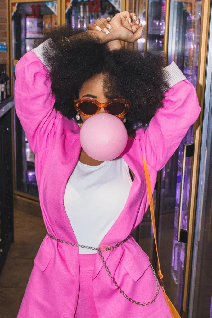 Person wearing a pink outfit and sunglasses, blowing a pink bubble gum bubble with arms raised