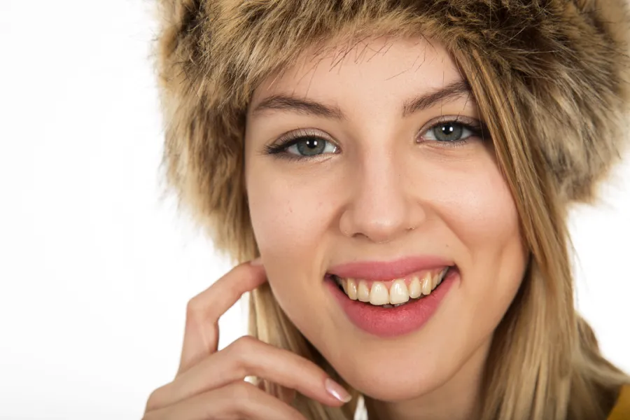 A person smiling, wearing a furry hat, with blonde hair visible and a finger touching their cheek